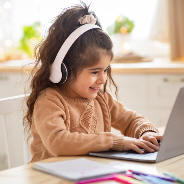 A little girl wearing headphones is using a laptop at home.