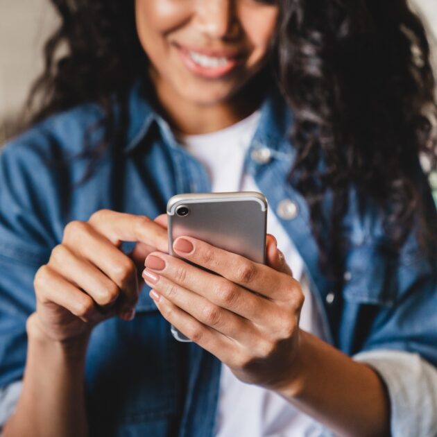 A woman in a denim jacket uses a smartphone, smiling and engaging with the device.